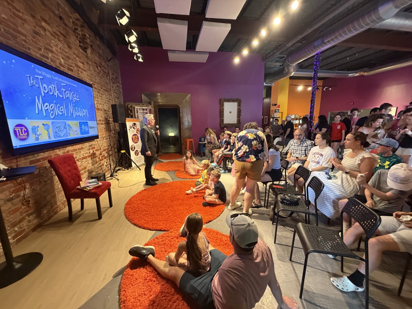 A full house at TLC Studios in Dedham watching the world premiere of The Tooth Fairy's Secret Workshop, with families seated on orange rugs and chairs in front of the film's title screen